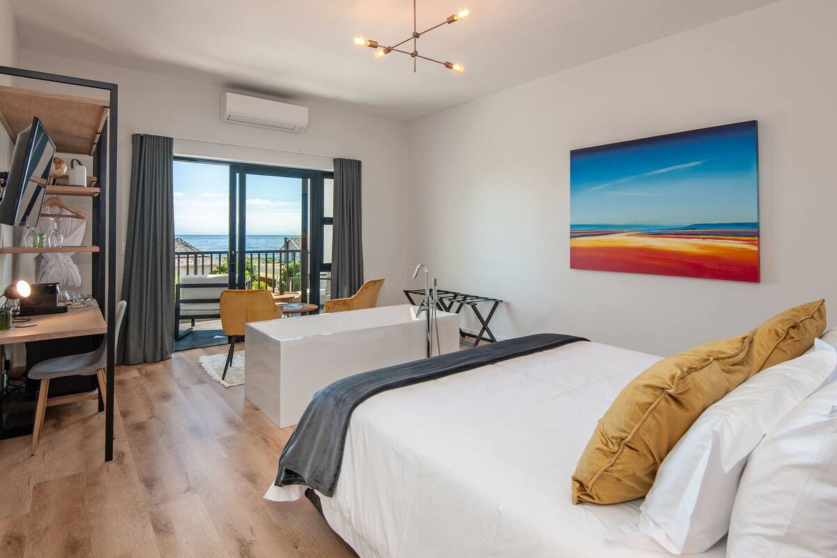 A modern hotel room with a large bed, a desk, and a balcony with a view of the sea.