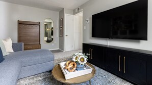 En moderne stue med en grå sofa, et sofabord med en bakke med snacks, et sort skab og et stort fladskærms-tv monteret på væggen.