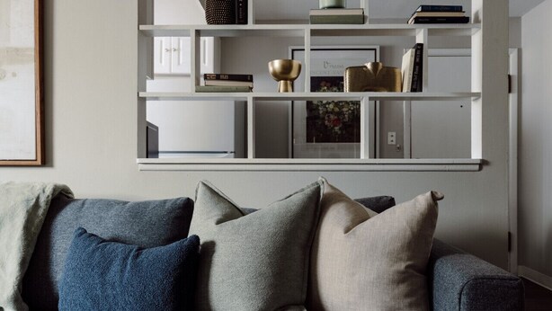 En grå sofa med flere puder i en stue med en hvid indbygget reol over.