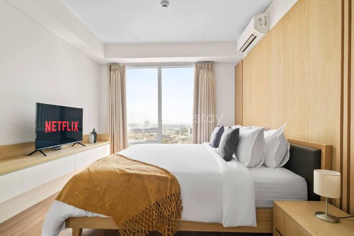 A hotel room with a bed, a TV displaying 'NETFLIX', a window with curtains, and a city view.