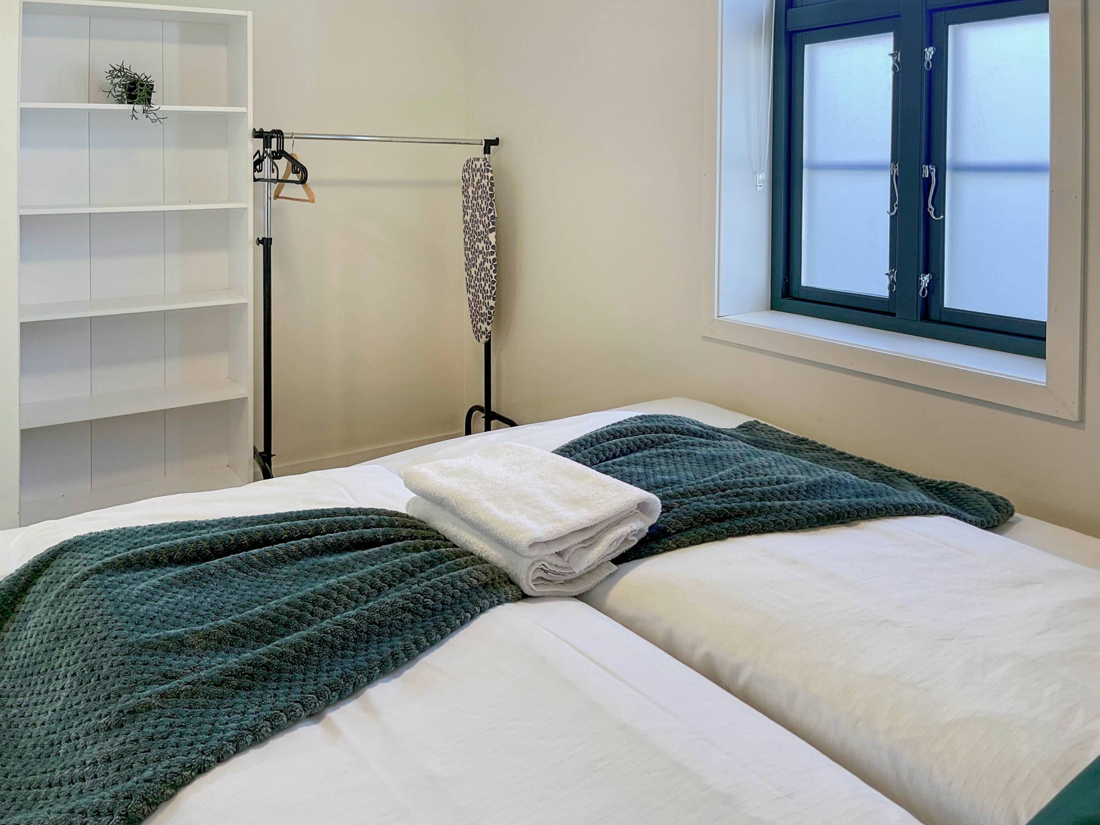 A neatly made bed with a green blanket, a white towel, and a shelf with a plant.