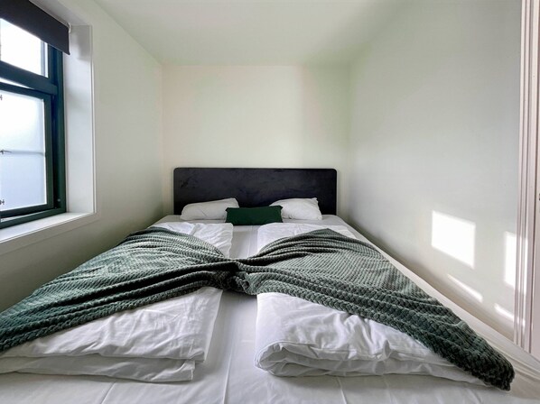A small, neatly made bed with white linens and a dark headboard, positioned in a room with white walls and a window on one side. - Vibrant Apartment on Bustling Street, Above a Restaurant - Perfect for Tourists, (Stavanger)