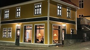 A two-story building with a yellow facade, large windows, and a visible dining area with a lamp. - Vibrant Apartment on Bustling Street, Above a Restaurant - Perfect for Tourists, (Stavanger)
