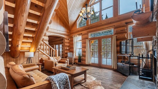 A log cabin interior with a staircase, leather sofas, a stone fireplace, and large windows. - Fiddler Lake Resort - Bear 10 (Mille-Isles)
