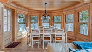 Interior de una cabaña de troncos con comedor, paredes de madera y ventanas grandes.