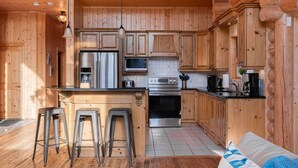 Interior de una cabaña de troncos con una cocina que incluye armarios de madera, un refrigerador de acero inoxidable, un microondas, una estufa y taburetes de bar.
