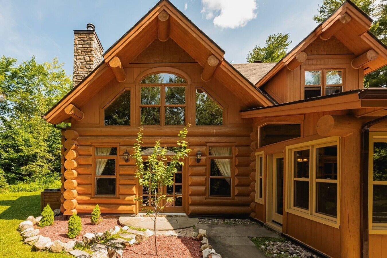Une cabane en rondins avec une cheminée, de grandes fenêtres et des fondations en pierre.