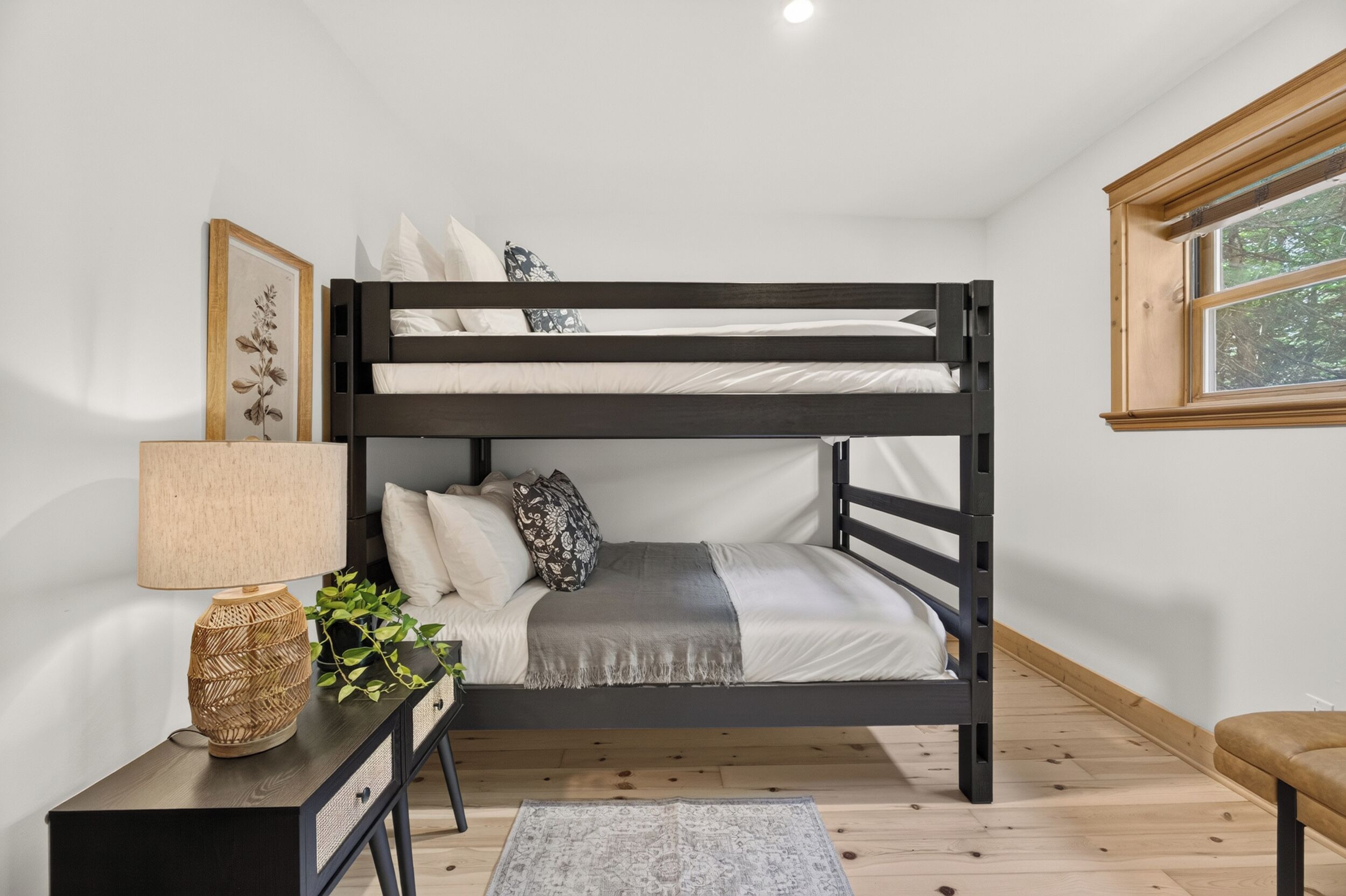 A bunk bed with a bedside table, lamp, and a window with a view of trees.