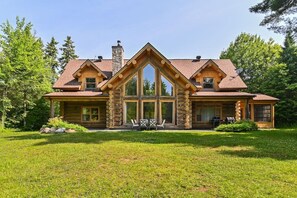 A log cabin with a stone chimney, large windows, and a grassy front yard.