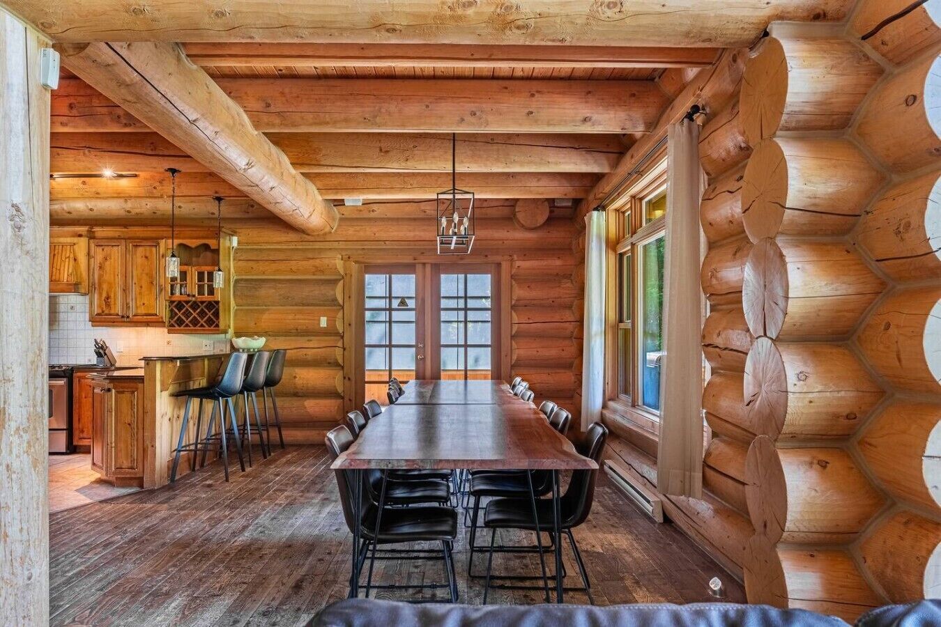 Interior de una cabaña de troncos con una larga mesa de comedor, sillas, una zona de cocina y amplias ventanas.