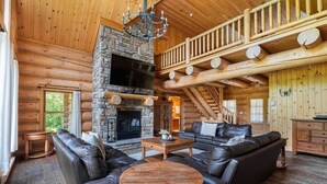 A log cabin interior with a stone fireplace, leather sofas, a wooden coffee table, and a wooden staircase.