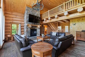 A log cabin interior with a stone fireplace, leather sofas, a wooden coffee table, and a wooden staircase.