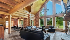 A log cabin interior with a large black leather sofa, dining area, and large windows offering a view of the outdoors.