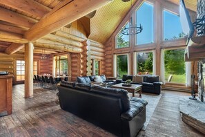 A log cabin interior with a large black leather sofa, dining area, and large windows offering a view of the outdoors.