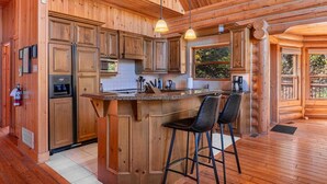 A wooden cabin interior with a kitchen featuring a central island, bar stools, and a large window.