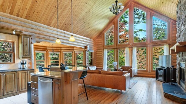 A spacious log cabin interior with a kitchen, dining area, and living room, featuring large windows and a fireplace.