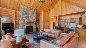 A log cabin interior with a stone fireplace, leather furniture, and a kitchen area.