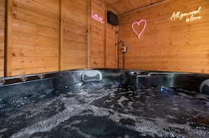 A wooden cabin with a hot tub, neon signs reading "love" and "All you need is love," and a heart-shaped neon light.