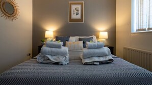 Cama bem arrumada com duas toalhas dobradas, um quadro na parede acima da cama e duas luminárias de cabeceira.