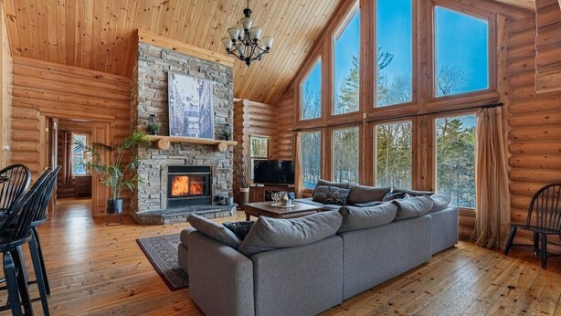 A log cabin interior with a stone fireplace, a gray sofa, and large windows.