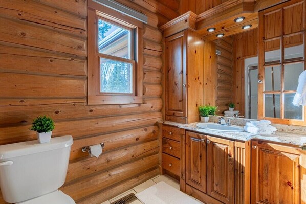 A log cabin bathroom with a toilet, sink, cabinets, and a window. - Fiddler Lake Resort - Deer 69 (Mille-Isles)
