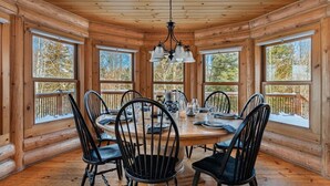 A wooden cabin interior with a round dining table, black chairs, and large windows.