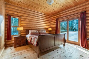 A log cabin bedroom with a wooden bed, nightstands, and a view of a snowy landscape.