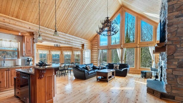 A log cabin interior with a dining area, living room with sofas, large windows, and a stone fireplace. - Fiddler Lake Resort - Deer 9 (Mille-Isles)
