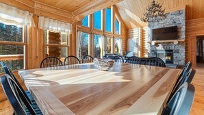 A log cabin interior with a large dining table, stone fireplace, and large windows. - Fiddler Lake Resort - Deer 9 (Mille-Isles)