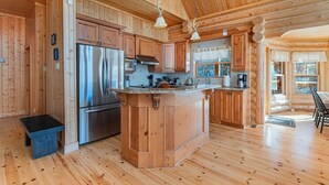 A log cabin interior with a kitchen featuring wooden cabinets, a stainless steel refrigerator, and a central island. - Fiddler Lake Resort - Deer 9 (Mille-Isles)