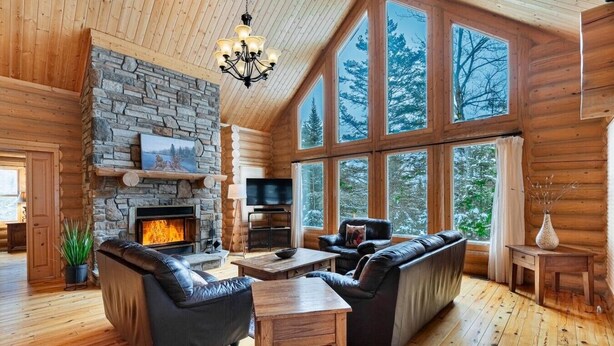 A log cabin interior with a stone fireplace, leather sofas, a wooden dining table, and large windows. - Fiddler Lake Resort - Deer 30 (Mille-Isles)