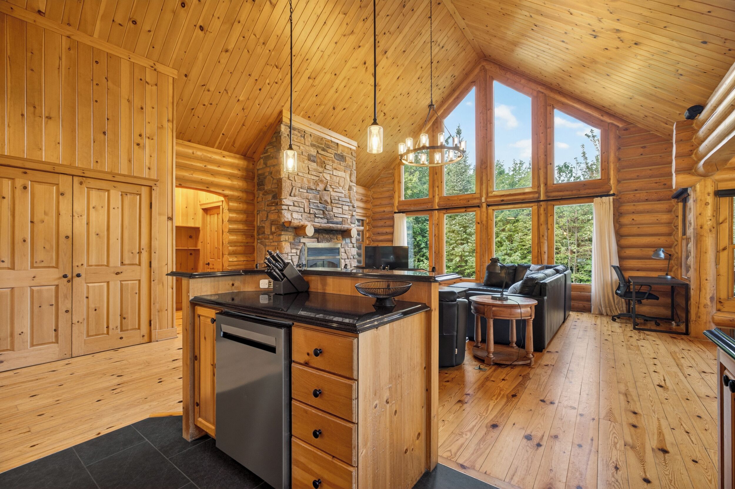 A log cabin interior with a kitchen, dining area, and a large window offering a view of the outdoors.