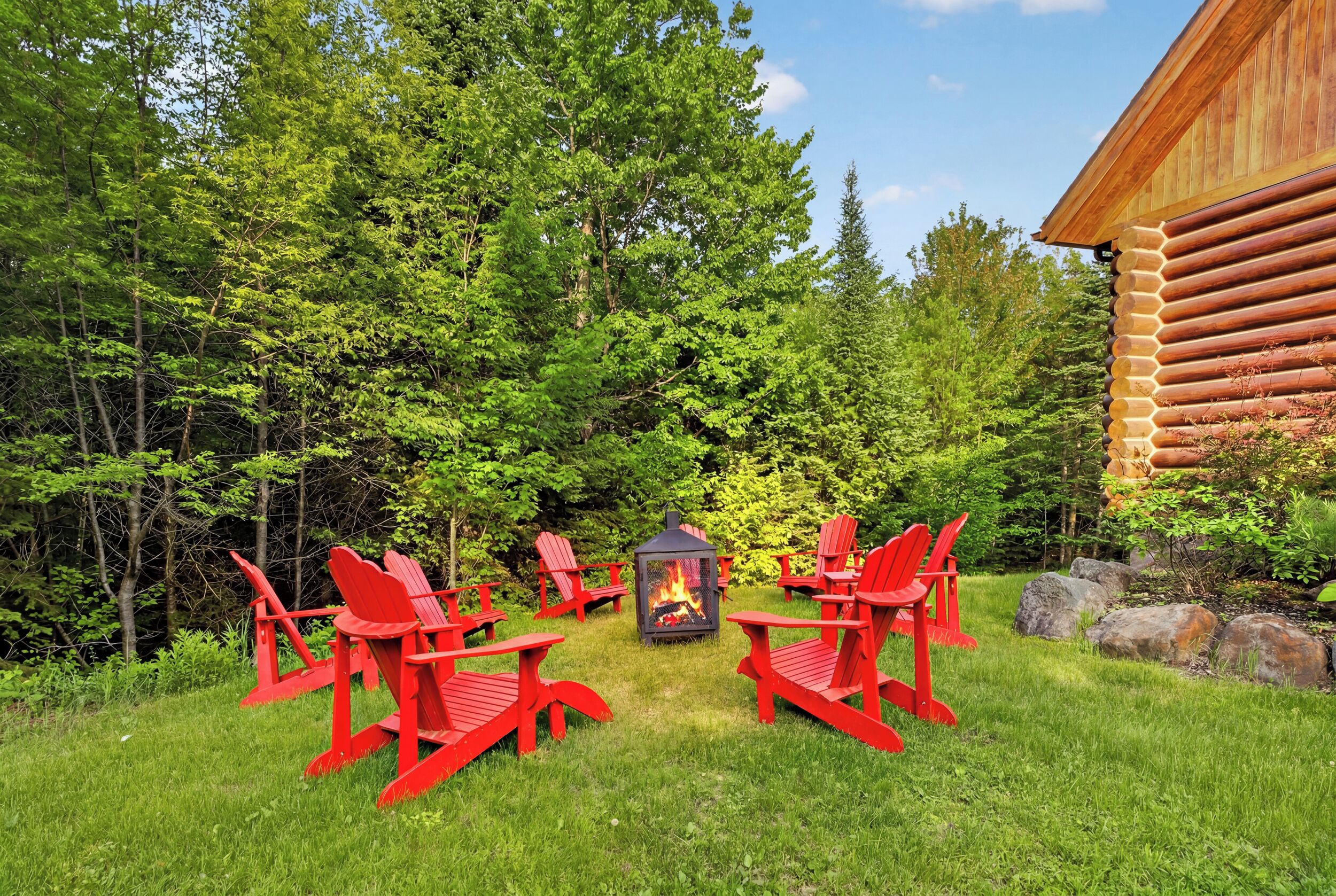 Une cabane en rondins avec un foyer et des chaises Adirondack rouges, située dans une zone boisée.