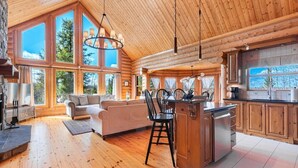 A spacious living area with a large sofa, dining table, and chairs, wooden floors, and large windows offering a view of the outdoors. - Fiddler Lake Resort - Deer 86 (Mille-Isles)