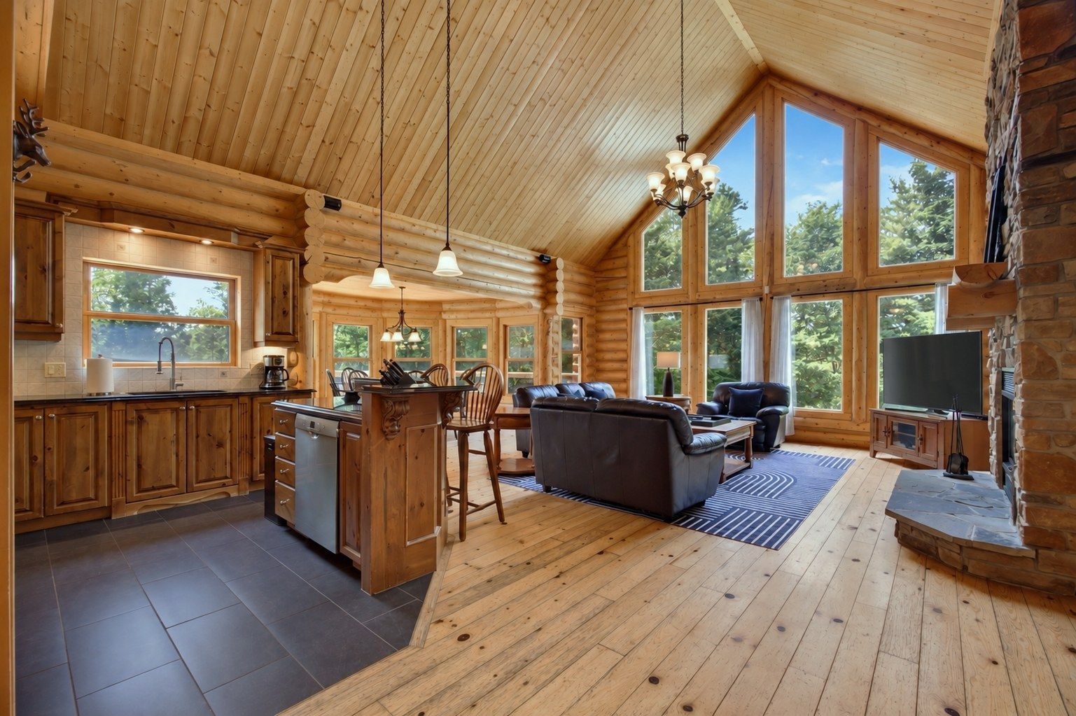 L’intérieur d’une cabane en rondins comprend une cuisine, un coin repas et un salon, avec de grandes fenêtres et une cheminée en pierre.