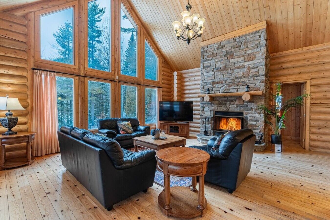 A log cabin interior with a stone fireplace, leather sofas, a TV, and large windows.
