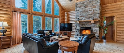 Interior de una cabaña de madera con chimenea de piedra, sofás de cuero, televisor y ventanas amplias.