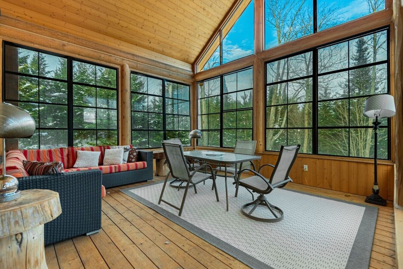 Un espacio exterior cubierto con piso de madera, un sofá, una mesa de comedor con sillas y ventanas amplias.
