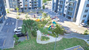 A multi-story residential building with a parking lot, a children's playground, and a landscaped area with palm trees. - Premium Space In Santiago- 5 Min Away From Airport (Puñal)