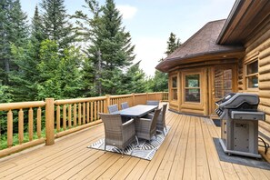 Eine Holzterrasse mit Tisch, Stühlen, einem Grill und Blick auf einen Wald.