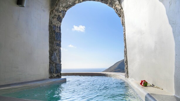 A stone archway leading to a pool with a clear view of the sea and a red rose on the edge. - Deep Earth Villas (Santorini)