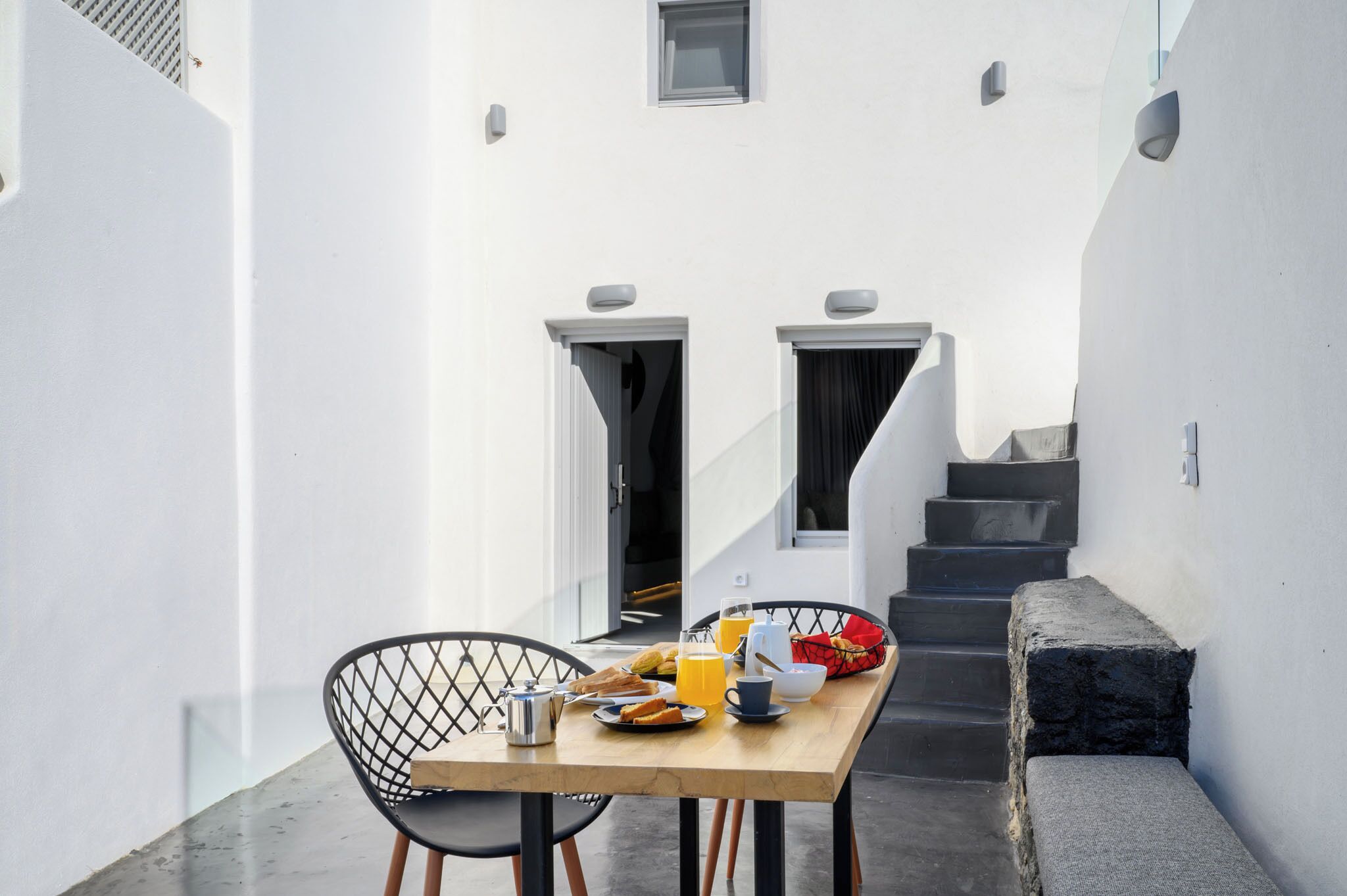 A narrow outdoor corridor with a wooden table set for breakfast, two chairs, and a staircase leading to an upper level.