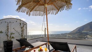 A rooftop terrace with two lounge chairs, a round table with fruits and drinks, a decorative umbrella, and a view of the city and hills. - Deep Earth Villas (Santorini)