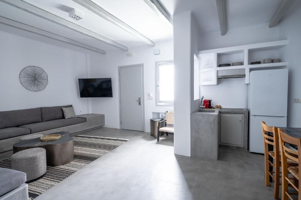 Un espacio habitable moderno con un sofá gris, un televisor de pantalla plana, una zona de cocina con armarios blancos y una mesa de comedor con sillas.