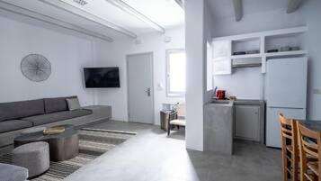 Un espacio habitable moderno con un sofá gris, un televisor de pantalla plana, una zona de cocina con armarios blancos y una mesa de comedor con sillas.