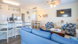 A living room with a blue sofa, two patterned armchairs, a dining table with chairs, a kitchen area with white cabinets, and a ceiling fan. - A Kure for the Sol (Kure Beach)