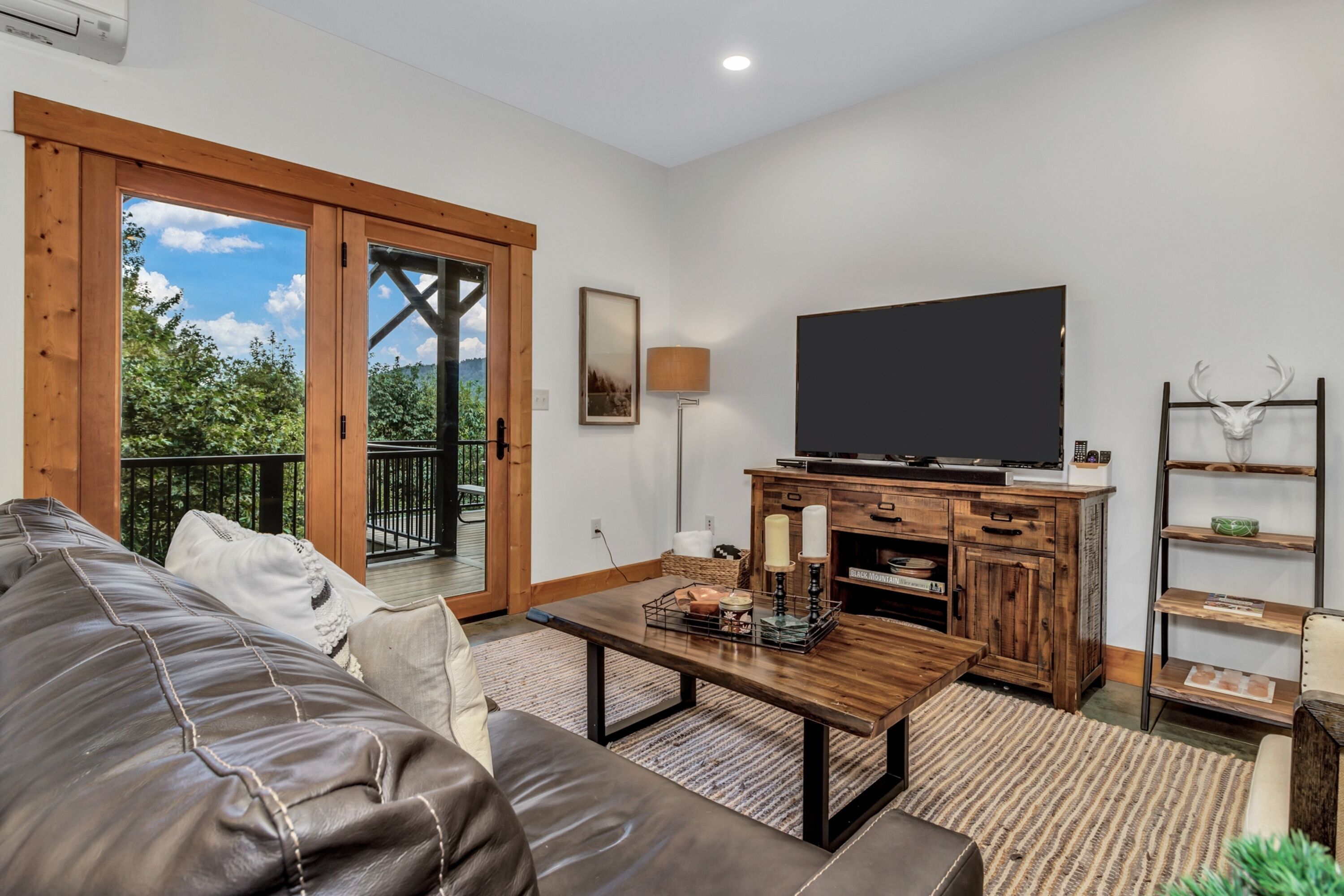 Ein Wohnzimmer mit Ledersofa, Couchtisch, Flachbildfernseher, Holzsärchen und einem Balkon mit Blick auf Grünflächen.