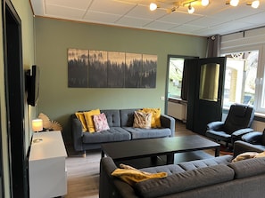 A modern living room with a grey sofa, a black coffee table, and a black armchair. There is a large window providing natural light and a view of the outside. - Platell Ferienhäuser Lerbach, Detached house for up to 8 people with fireplace (Osterode am Harz)