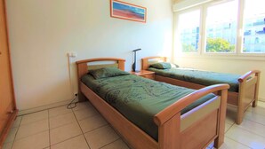 A room with two wooden beds, a nightstand, a lamp, a painting on the wall, and a window with a view of buildings. - Appart T3 Tout Confort + Parking Privatif (La Rochelle)