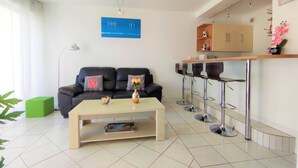 A modern living room with a black sofa, a coffee table, and a kitchen area with bar stools. - Appart T3 Tout Confort + Parking Privatif (La Rochelle)